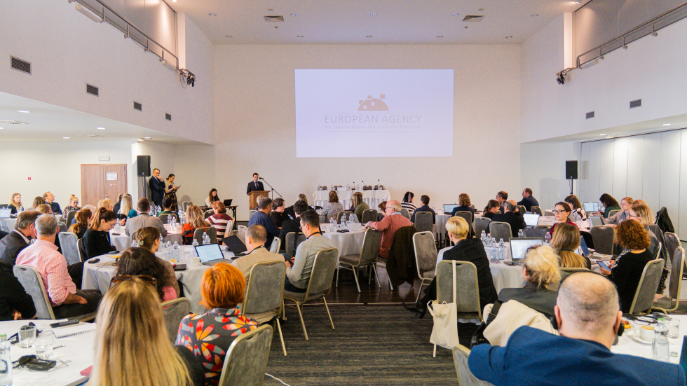 Participants at the bi-annual meeting sit around round tables and watch a presentation on a screen at the front of the room