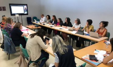 A group of people sit around a conference table, watching a presentation on a screen
