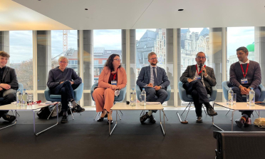 Six people sit on a stage, João Costa is holding a microphone and speaking 