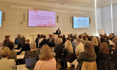A person presenting at the front of a room full of audience members sitting on chairs