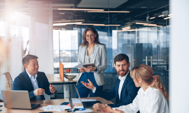 Three people sit at a conference table talking. A fourth person stands by the table, listening