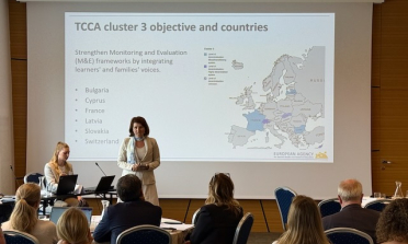 A person presenting at the front of a room full of audience members sitting on chairs. The screen behind the speaker says 'TCCA cluster 3 objective and countries'